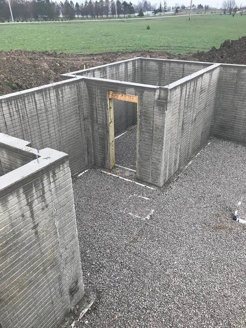 Concrete foundation with open door, wooden framing, partial brick wall, grassy yard bordered by trees and fence, clear sky overhead.