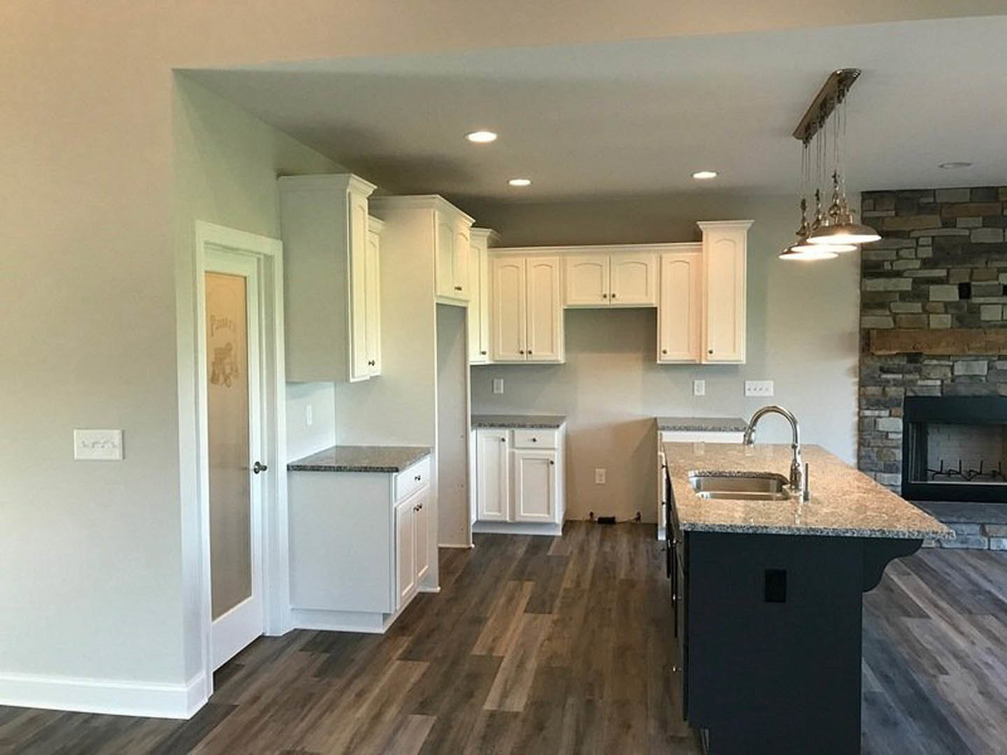Kitchen with stone fireplace, white cabinetry with black knobs, white countertop, kitchen island with built-in sink, chandelier overhead, tiled backsplash, stainless steel