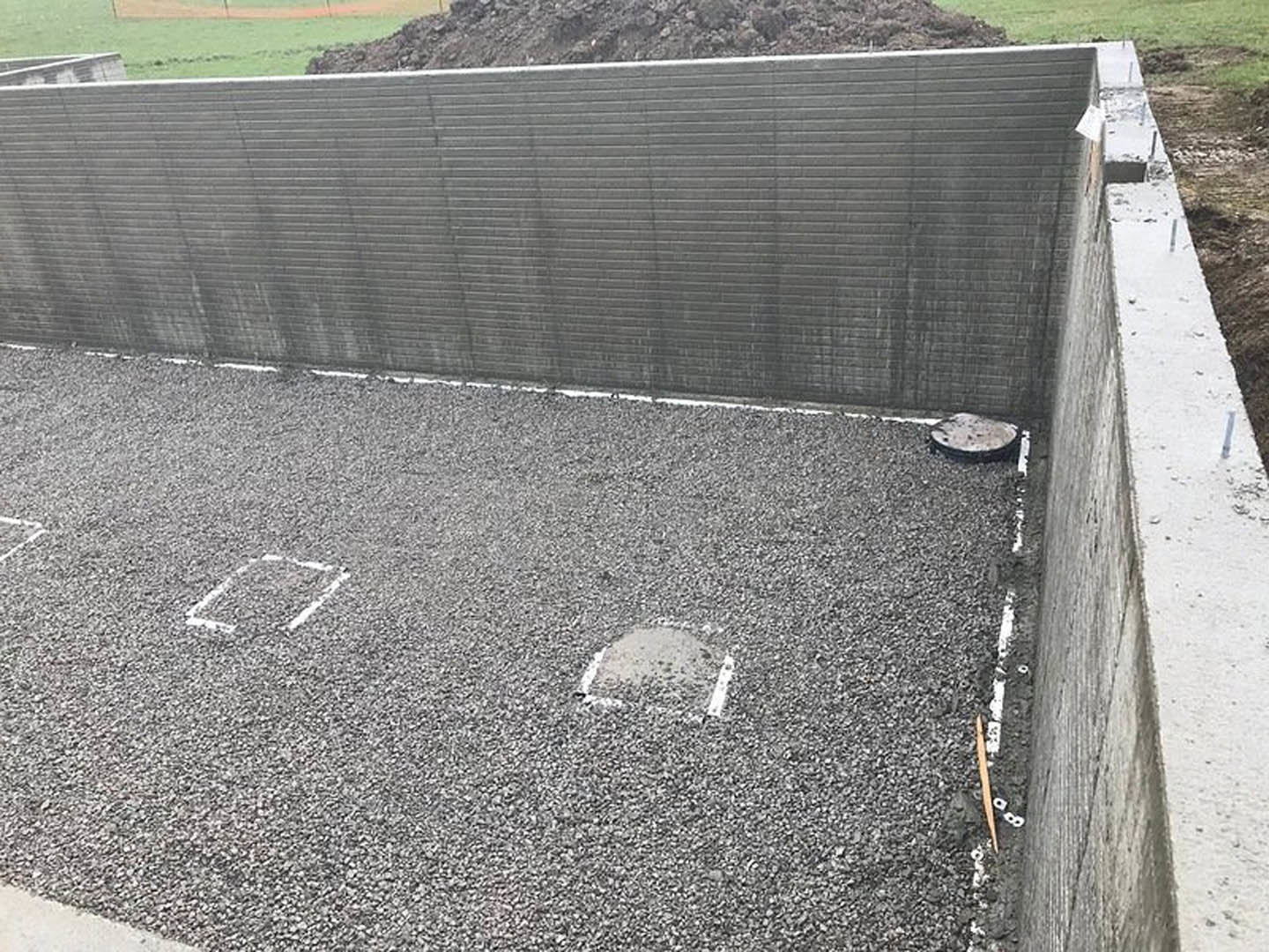 Grey concrete wall with square openings, adjacent to a cement wall and dirt-covered brick wall; white bucket and pole in foreground, sparse grass and snow visible.
