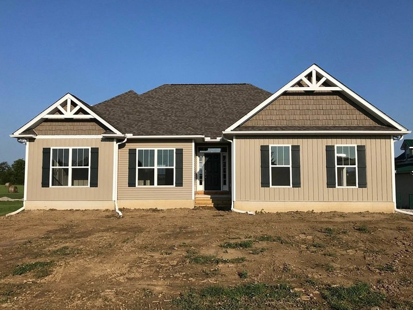 Two-story house under construction with black roof, white-framed windows, and light siding, surrounded by dirt and patches of grass under clear blue sky