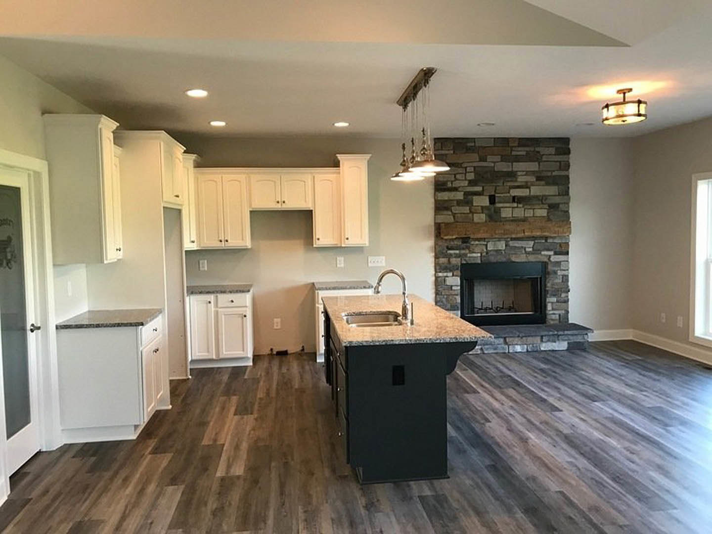 Kitchen with white cabinets and black knobs, stone fireplace, central island with sink, wood and laminate flooring, chandelier overhead, close-up of faucet and fireplace details