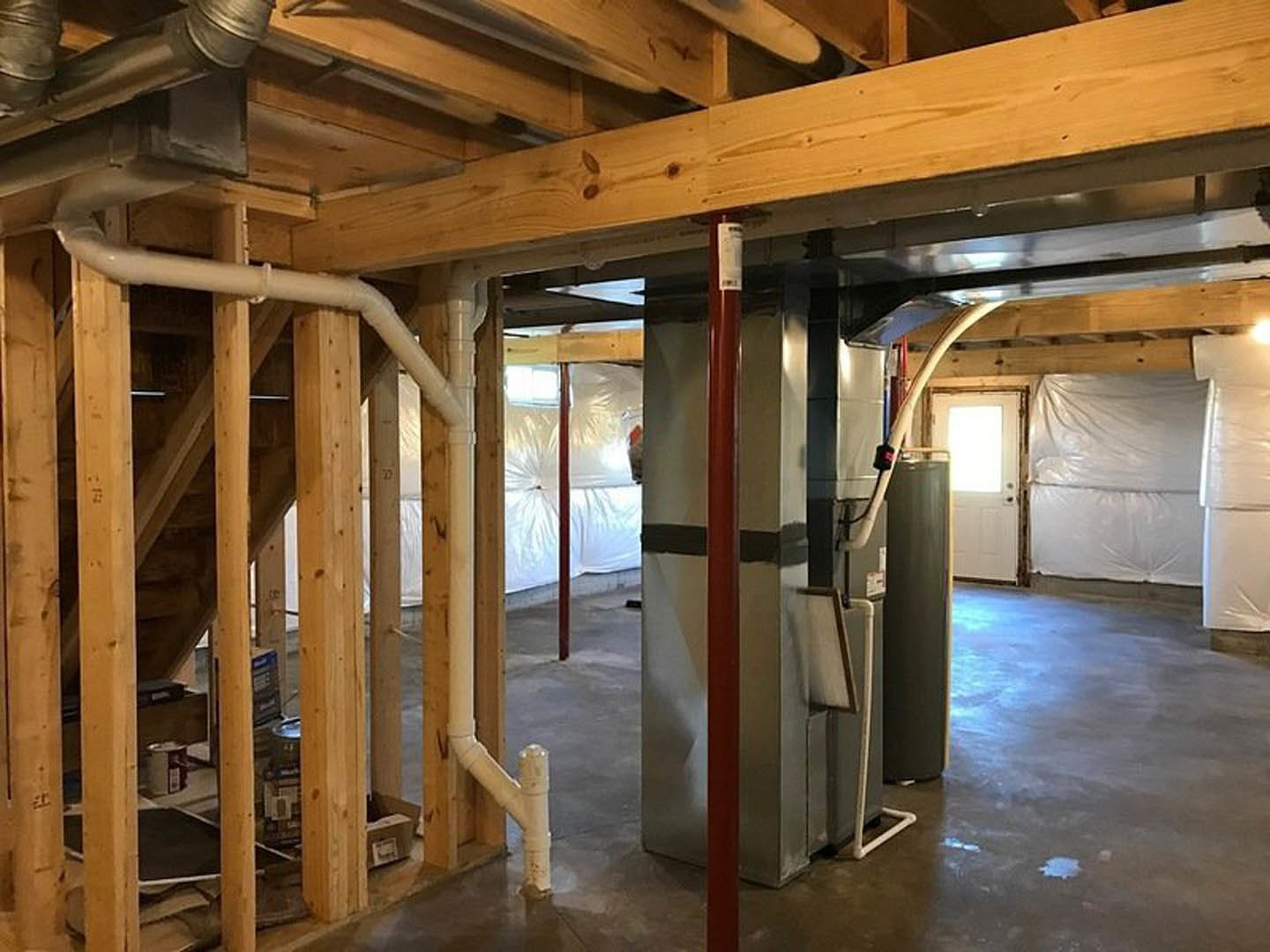 Unfinished room with exposed pipes along ceiling and walls, white plastic vapor barrier on wall, white door with light shining through, concrete floor, pillar, and construction