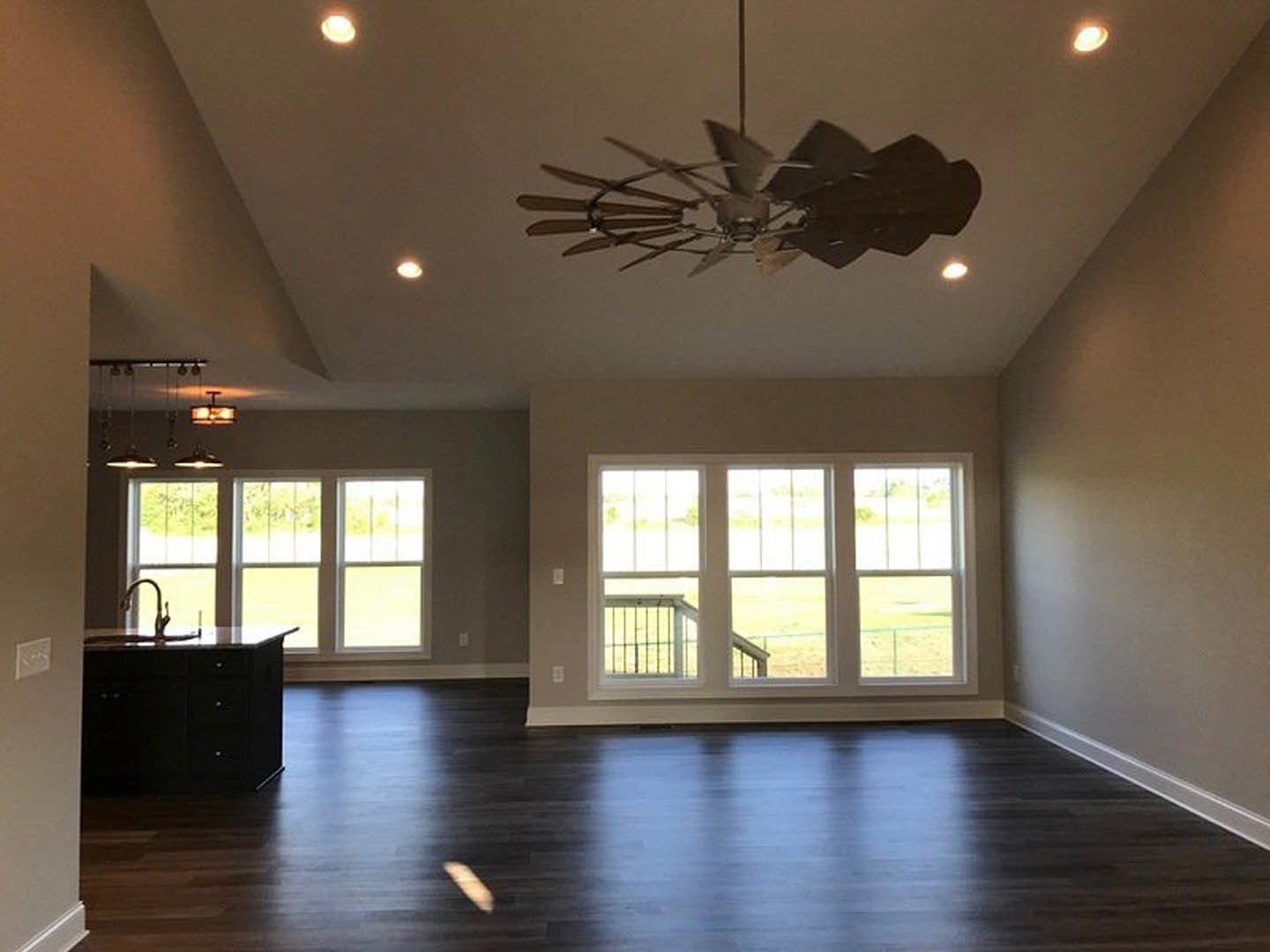 Open-concept room featuring dark wood flooring, white walls, ceiling fan with dark blades, large window with metal railing, black kitchen island with drawers, and white electrical