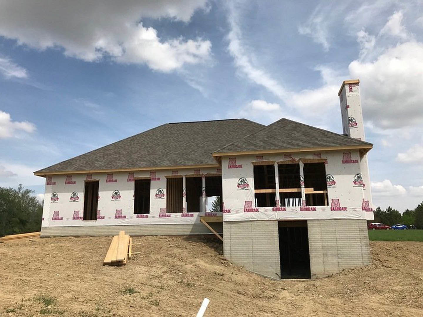 Partially built house with exposed framing and red tape, situated on a dirt hill under a clear blue sky, scattered construction materials including a wooden sled and white pipe in