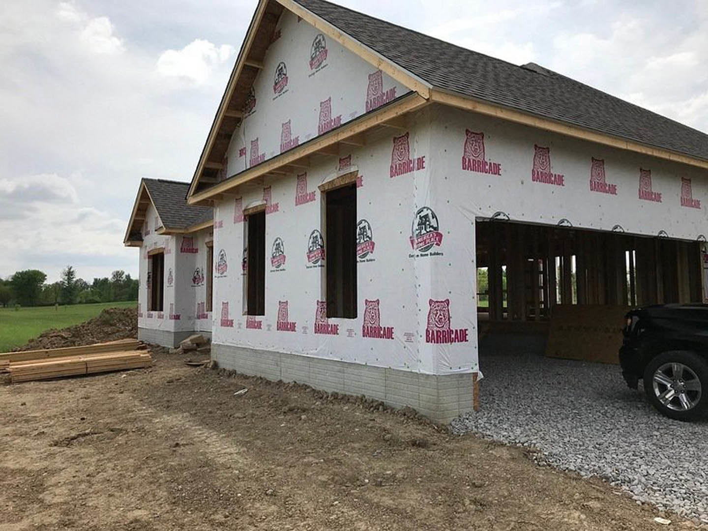 Partially built house with exposed wood framing, attached garage, gravel driveway, black car parked outside, construction materials scattered nearby