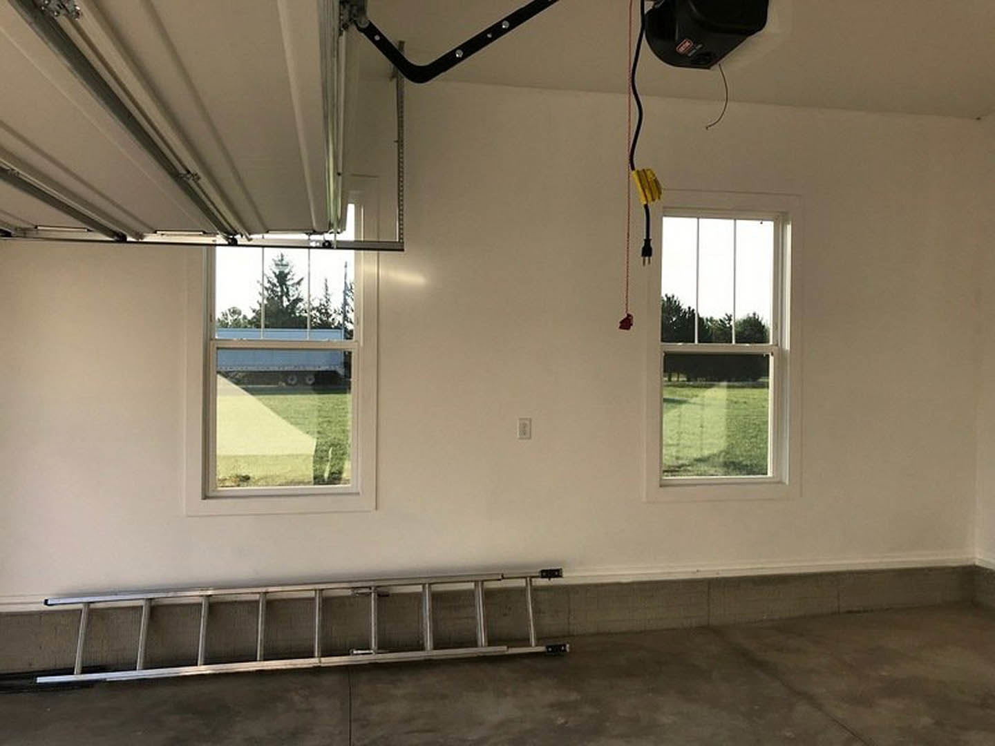 Metal ladder with curved handle leaning against plaster wall in concrete garage, window showing green lawn and parked trailer, black hat resting nearby on floor.