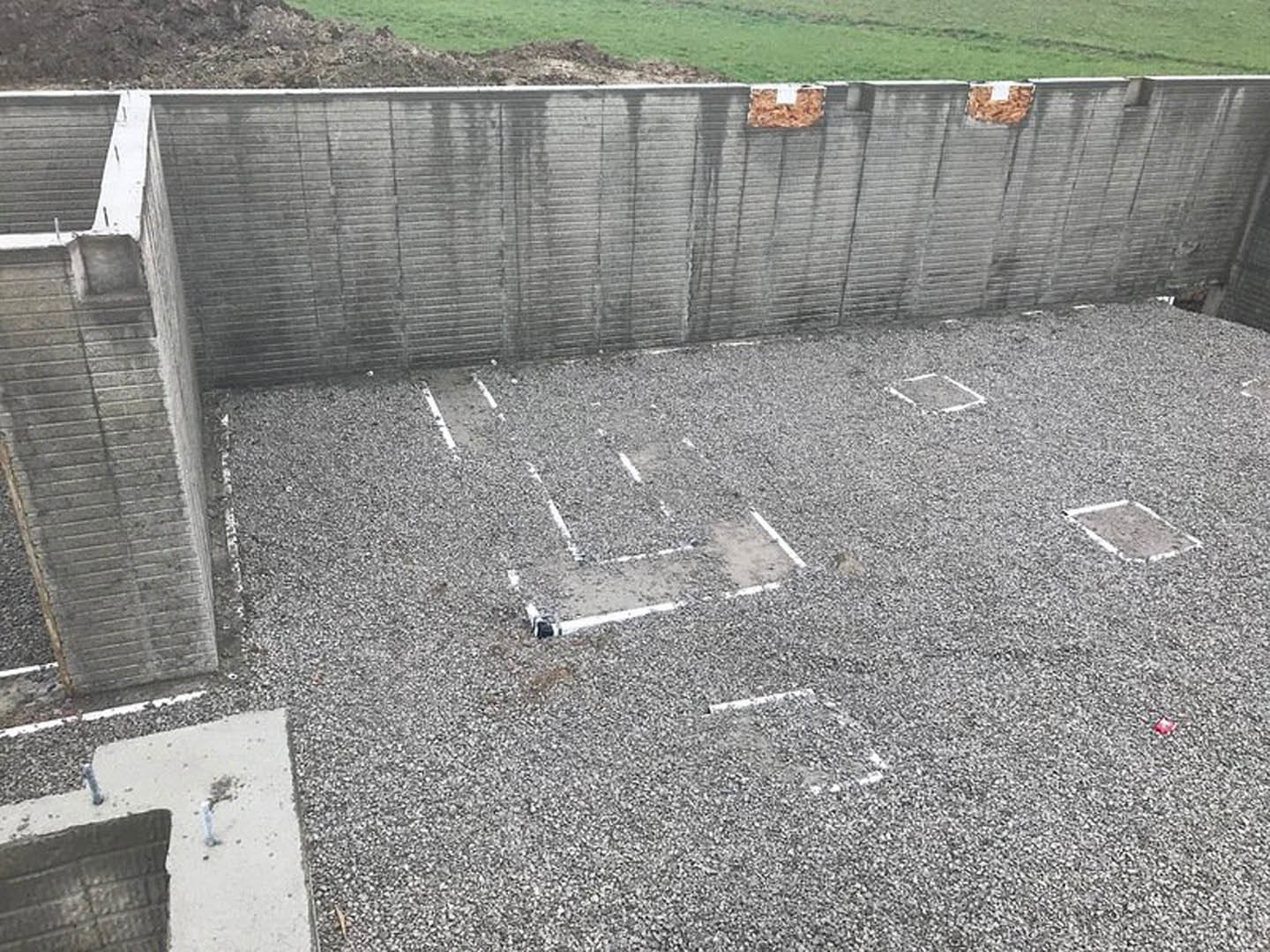 Modern home exterior featuring smooth concrete walls, a concrete slab foundation with visible bolts, grassy lawn, and a wood pile in a basket near the entrance.