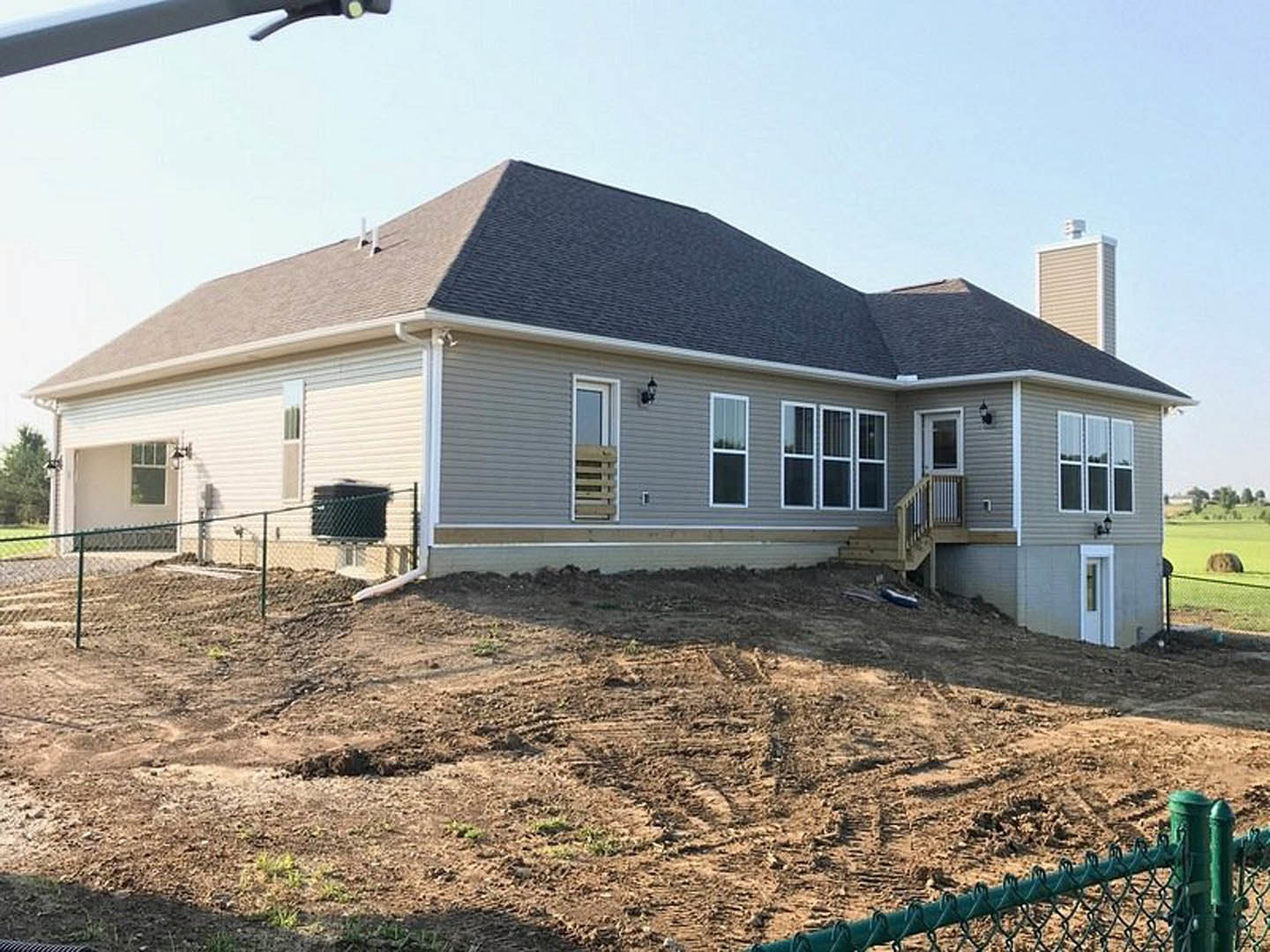 Two-story house under construction with exposed framing, white window frames, dirt yard with tire tracks, wooden fence in foreground