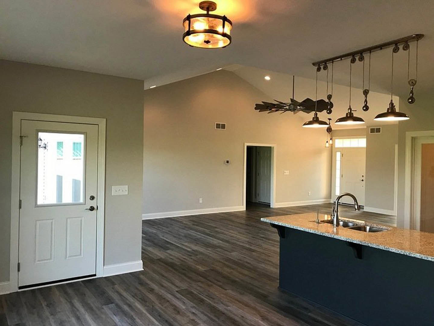 Open kitchen and dining area with wood flooring, white cabinetry, kitchen island with sink, glass-shaded ceiling light fixtures, white door with window and nearby light switch