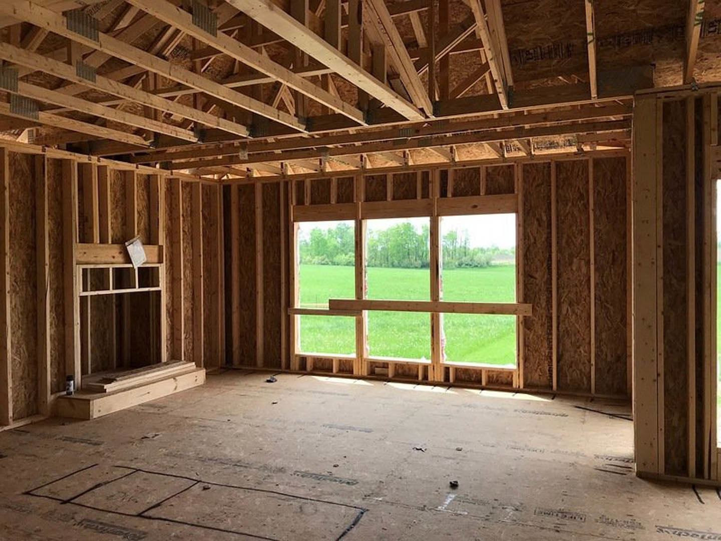 Sunlit room with wood plank flooring, white accent on wooden surface, large window framed in wood overlooking grassy field and trees.