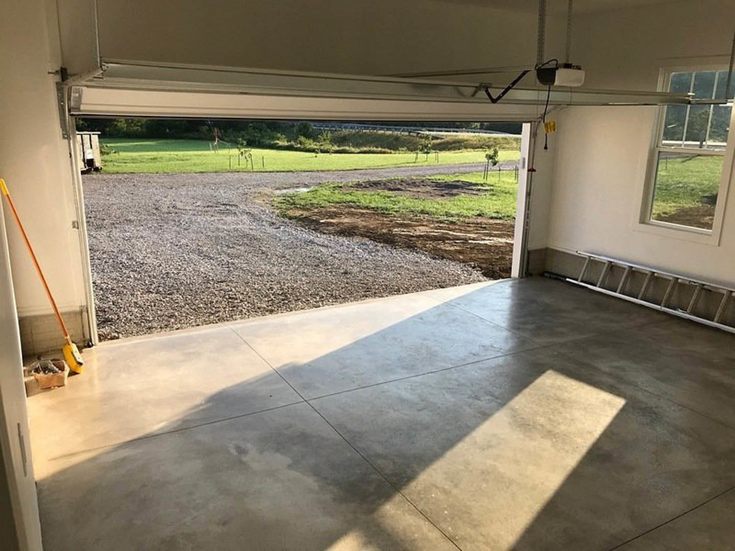 Garage interior with concrete flooring, gravel walkway, white wall featuring a long orange pencil, ladder propped against wall, window overlooking grassy field, gravel road