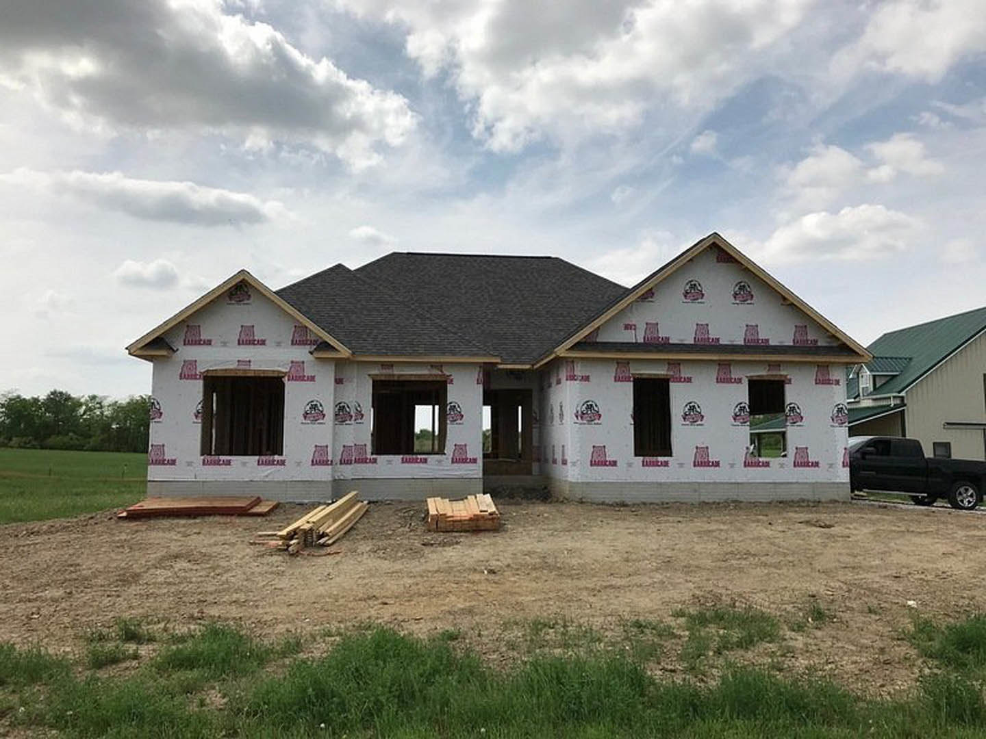 Framed house under construction with exposed plywood sheathing, red stickers on exterior walls, black pickup truck parked nearby, pile of lumber on grass, cloudy sky overhead