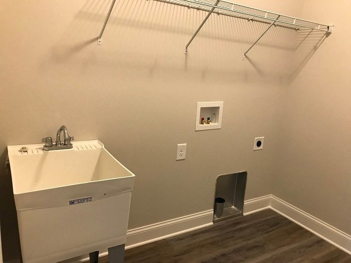Laundry room with white sink and silver faucet, small white square shelf, metal trash can in corner, electrical outlet, black drain cover on white wall