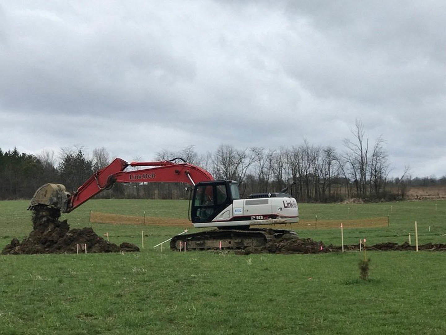 Bulldozer excavating earth in grassy field under cloudy sky, surrounded by trees and plants