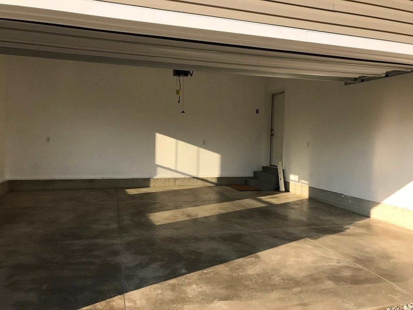 Empty garage interior with smooth concrete floor, white plaster walls, open door, ceiling lighting, electrical outlet, and window letting in natural light.
