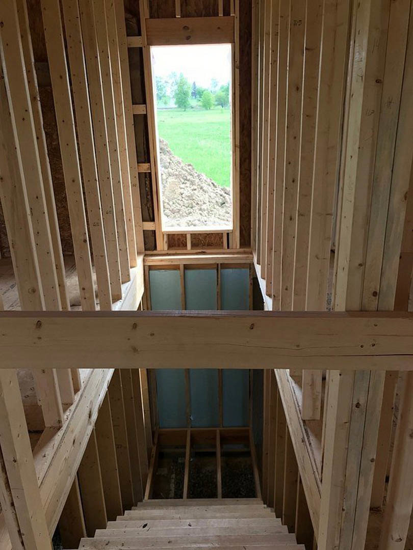 Unfinished room with exposed wood beams, partially installed insulation, large window overlooking a field, and wooden staircase beside a blue accent wall