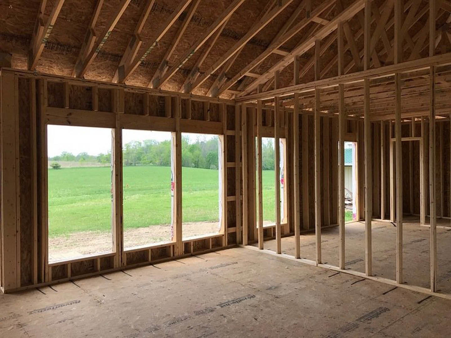 Wood-framed house under construction with exposed roof trusses, unfinished floors, large windows, and surrounding grass and trees