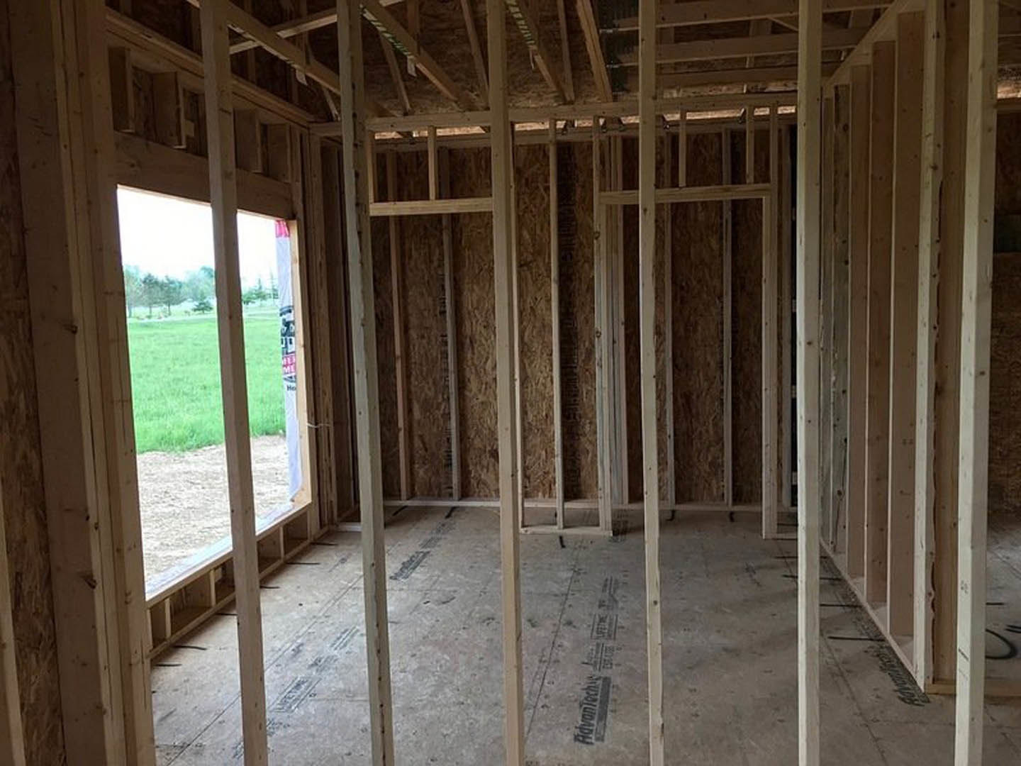 Room under construction with exposed wood framing, large window overlooking grassy yard, unfinished floor with wooden beams and planks, insulation visible between studs