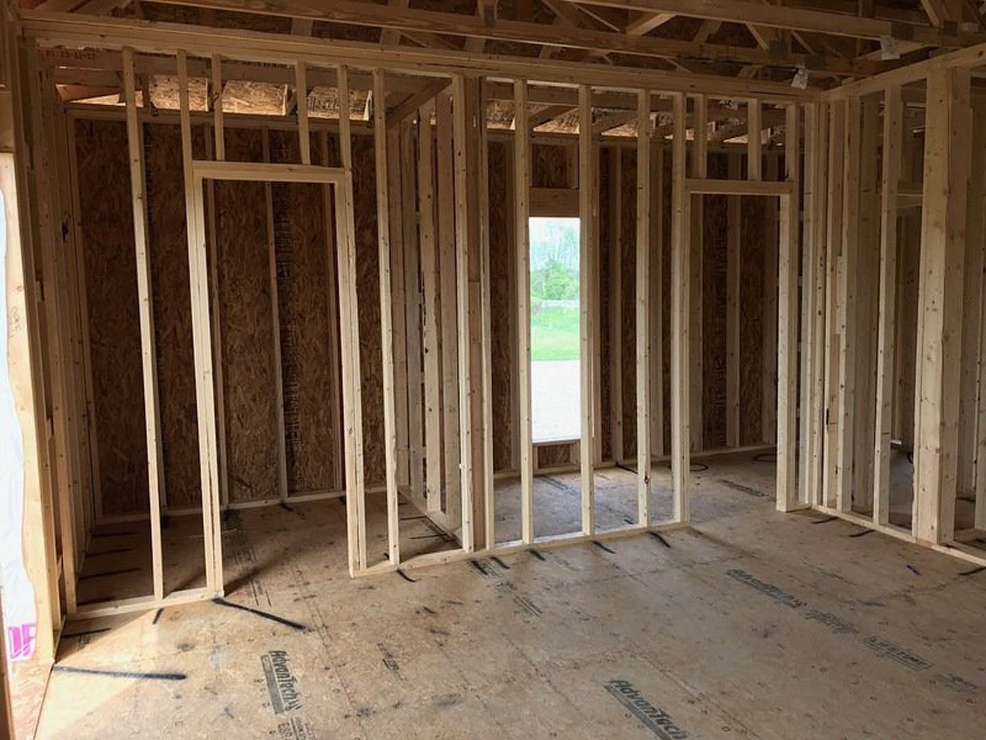 Wood-framed room with exposed ceiling beams, unfinished floor framing, large window overlooking green field, close-up of a sign and door visible
