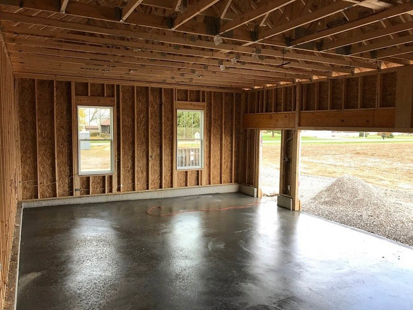Concrete floor with a red cord, large windows showing trees and yard, exposed wooden ceiling beams, pile of gravel beside sidewalk