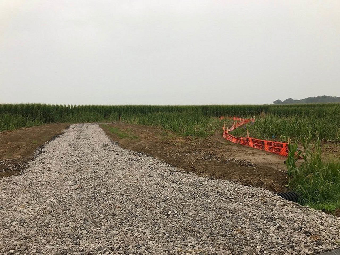 Gravel driveway bordered by orange barriers and lush green plants, with a fence running alongside grassy field under cloudy white sky