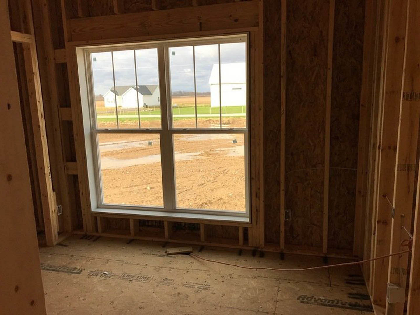 Large wood-framed window in a bright room with plank flooring, offering a view of a field, farm, and house behind a fence.