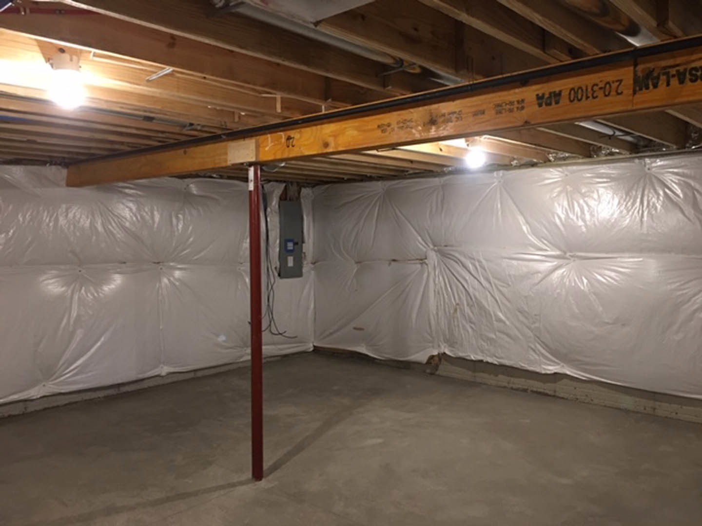 Basement with white plastered wall, exposed wooden ceiling beams, concrete floor, and bright overhead lighting