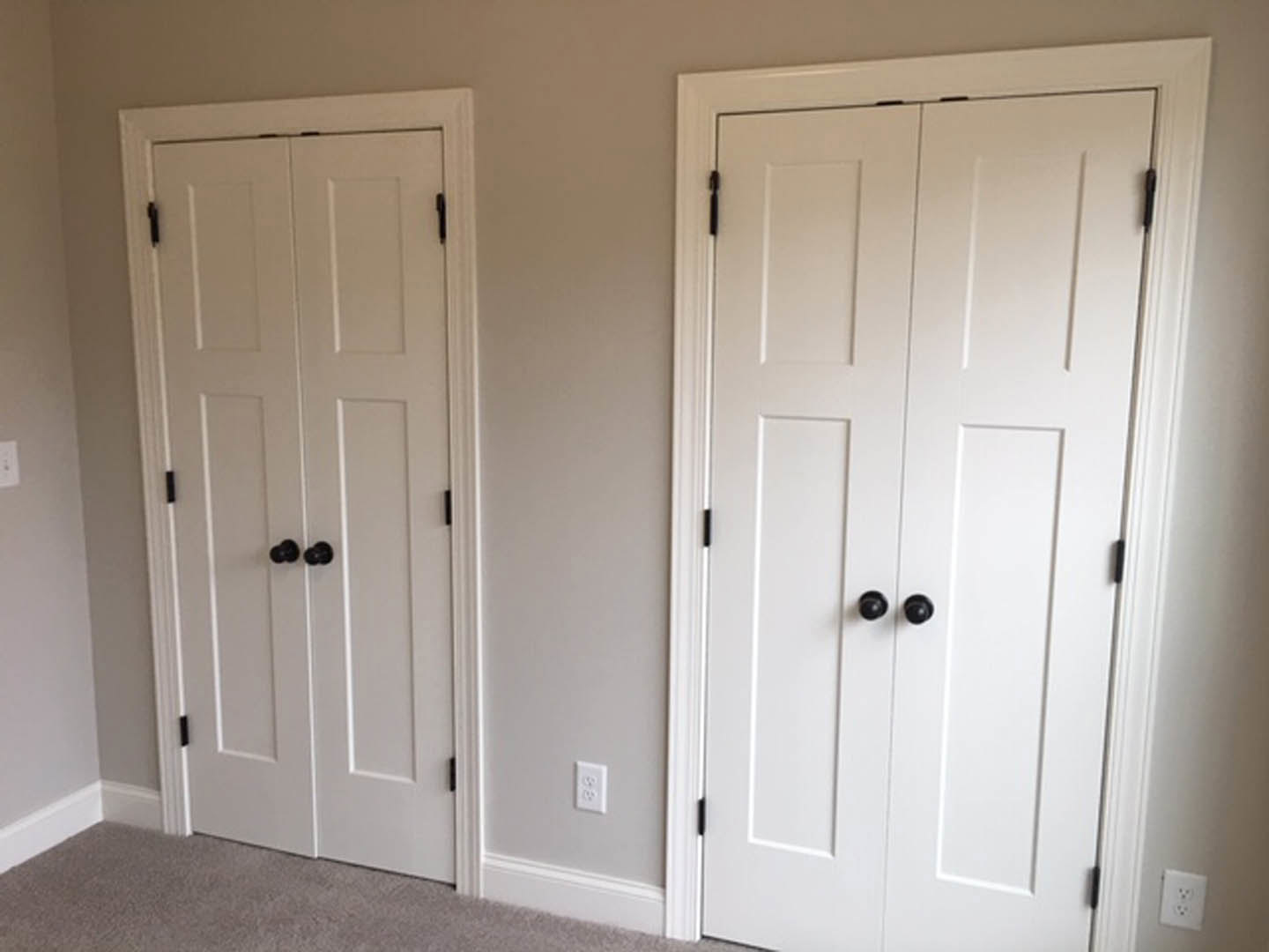 White paneled closet and entry doors with black knobs, white baseboards, light gray walls, and a white electrical outlet on hardwood flooring.