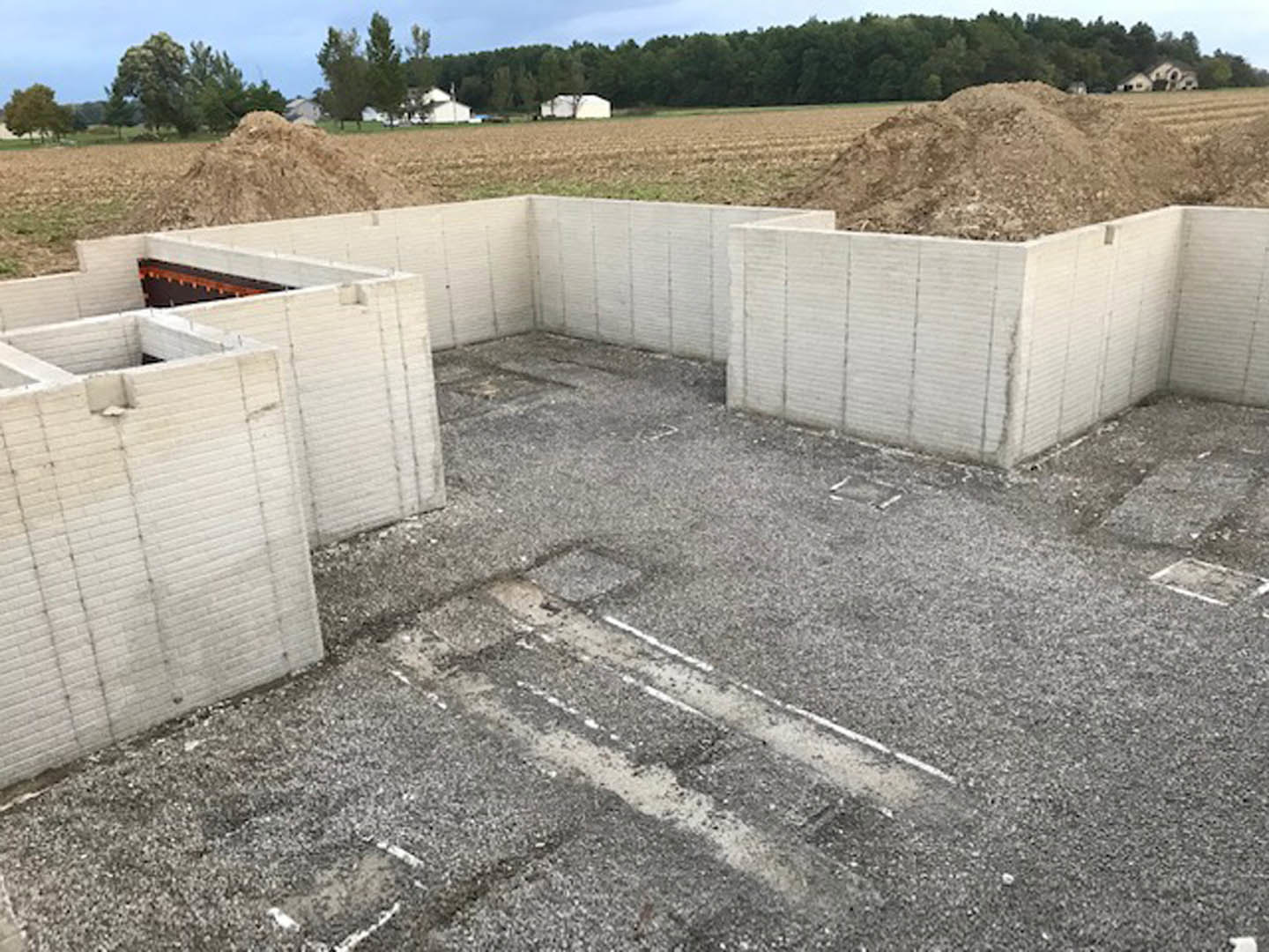 Construction site with exposed foundation, concrete walls, piles of dirt, scattered building materials, and surrounding trees under a clear sky
