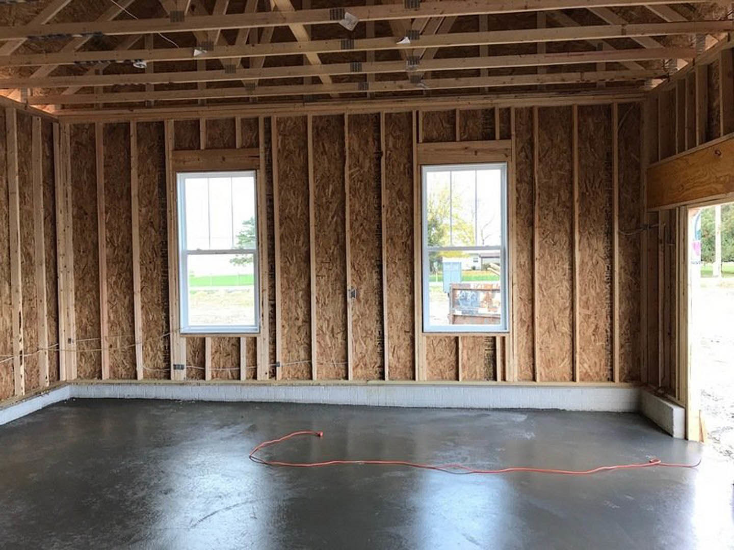 Spacious room featuring large white-framed windows, exposed wooden ceiling beams, concrete floor with a red cable, and view of a grassy yard