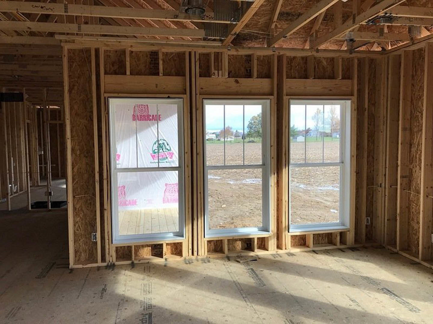 Spacious room featuring exposed wood ceiling beams, multiple large windows with plastic coverings, unfinished wood framing, and a view of an open field outside