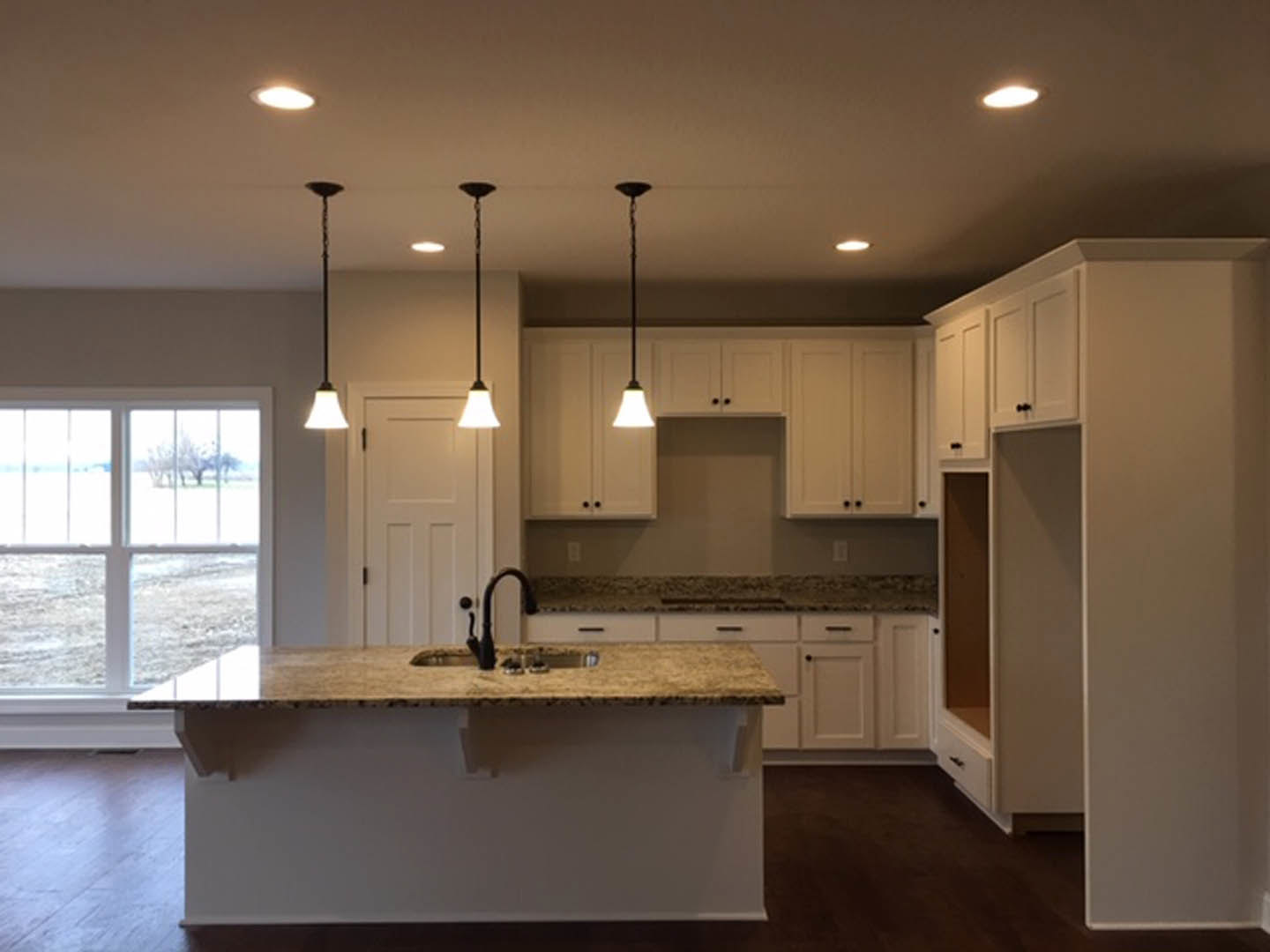 Modern kitchen featuring a central island with pendant lights, white tile backsplash, wood cabinetry, stainless steel sink and faucet, large window overlooking a grassy field, and