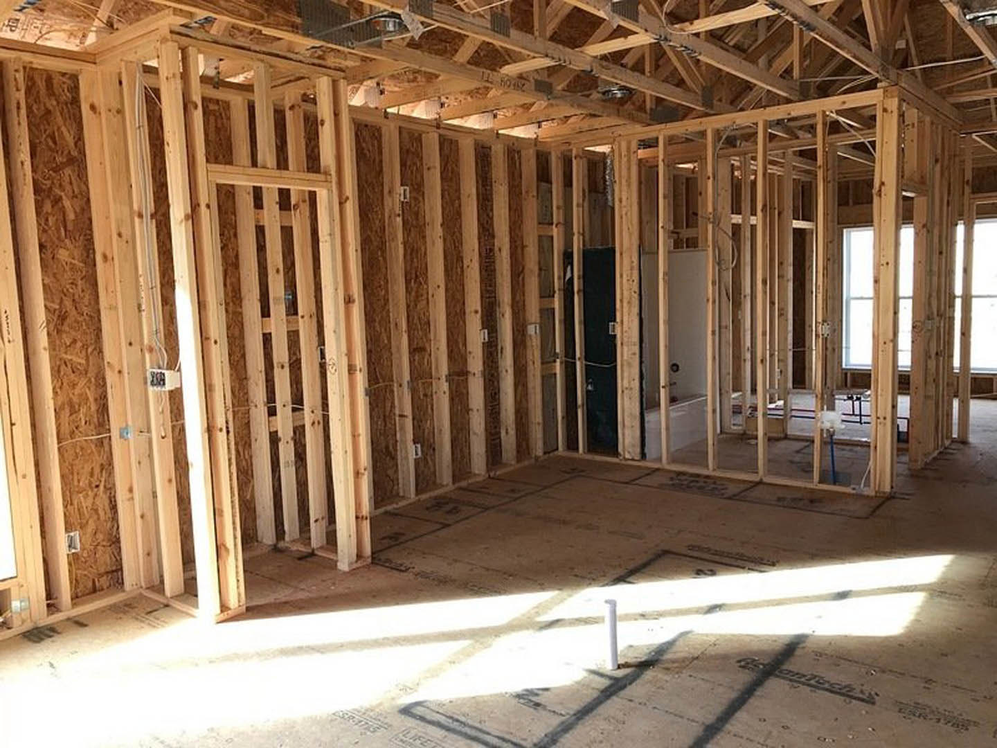 Wood-framed room under construction with exposed beams, unfinished walls, white pipe embedded in concrete floor, and doorway opening