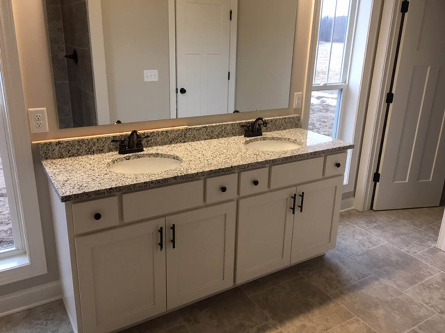 Double vanity bathroom with speckled granite countertop, large wall mirror, chrome faucets, white cabinetry, and a white door adjacent to tiled walls.