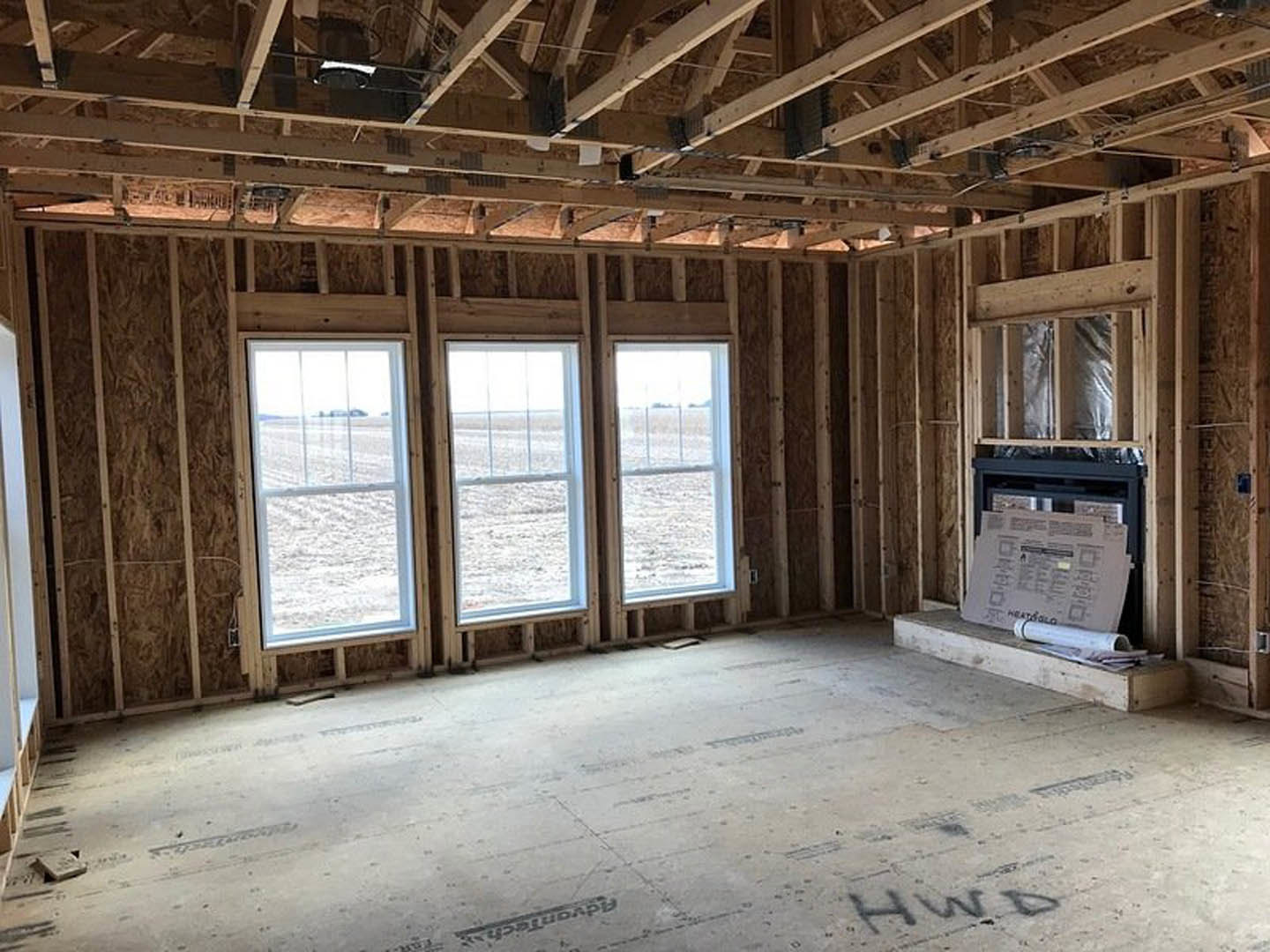 Living room with exposed wood beams, concrete floor, row of large windows overlooking a field, fireplace with stone surround, newspaper resting on window sill.