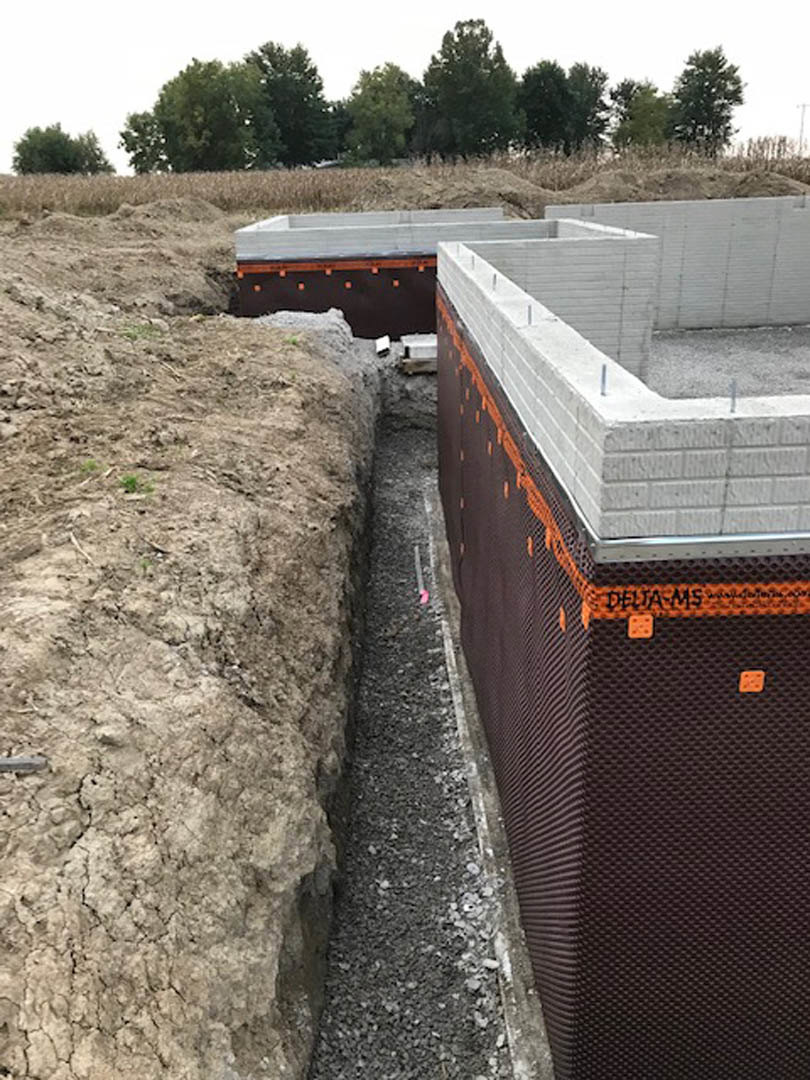 Concrete house foundation with exposed composite material, surrounded by dirt and construction track, trees and sky in background