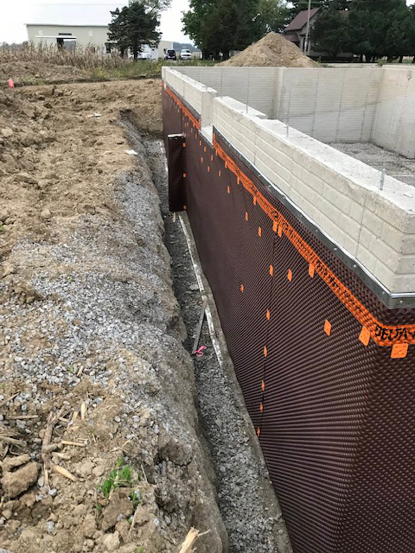 Concrete foundation with exposed orange and black plastic sheeting, adjacent piles of dirt and rocks, partially constructed walls with brown, white, and composite materials