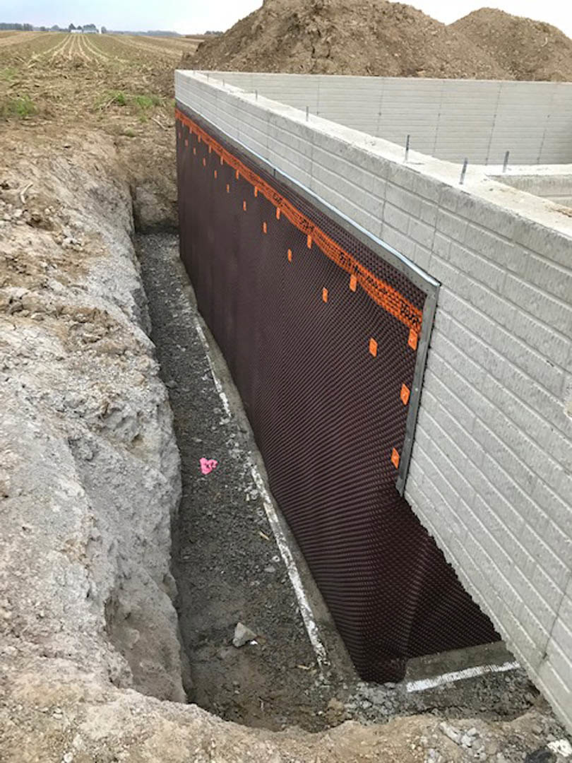 Concrete foundation wall with exposed metal rods, orange and black mesh along the base, dirt and rocks in the foreground, open sky above