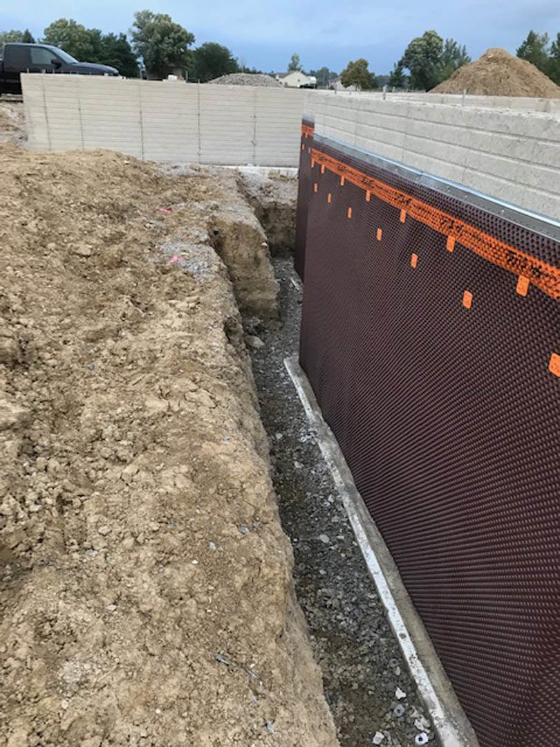 White exterior wall with a rectangular hole in the dirt ground, car parked in the background, tree partially visible, outdoor setting under clear sky.