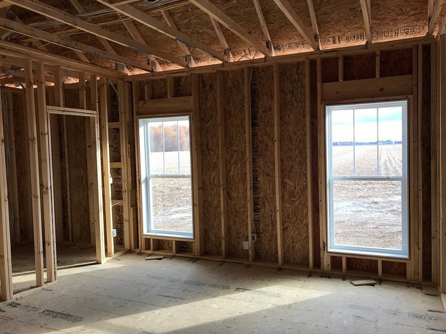 Open-concept room with large windows overlooking grassy field, exposed wood ceiling beams, light hardwood floors, and neutral walls