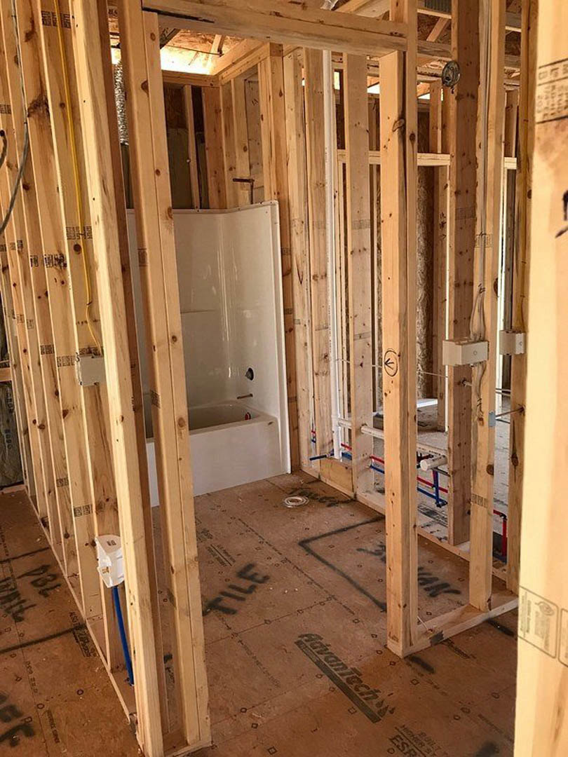 Bathroom under construction with exposed wood framing, blue plumbing pipe, unfinished wood floor marked with black text, open shower door, and construction materials including a
