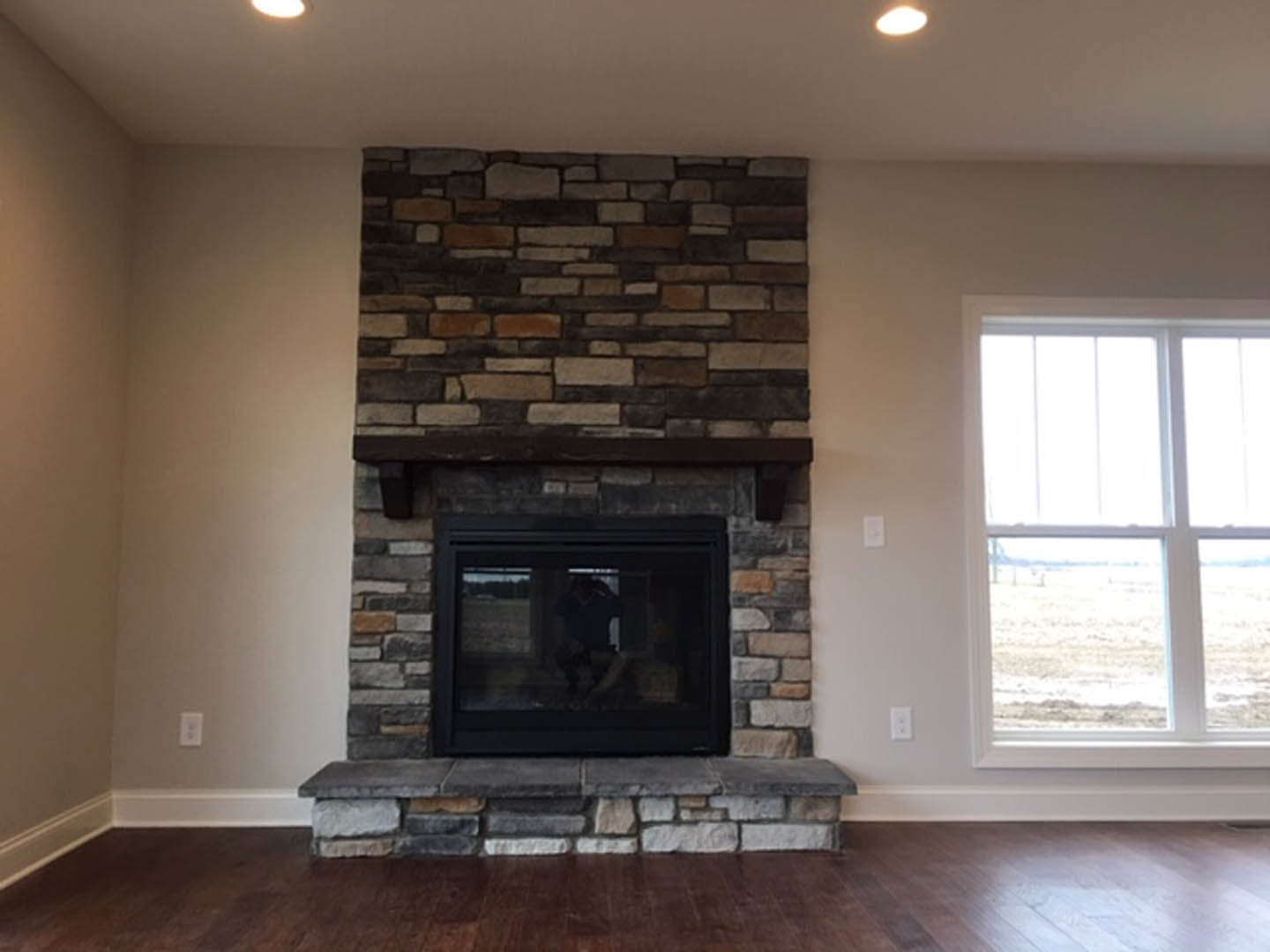Brick fireplace with wooden mantel in living room, hardwood floors, large window overlooking grassy field, built-in shelves along wall.