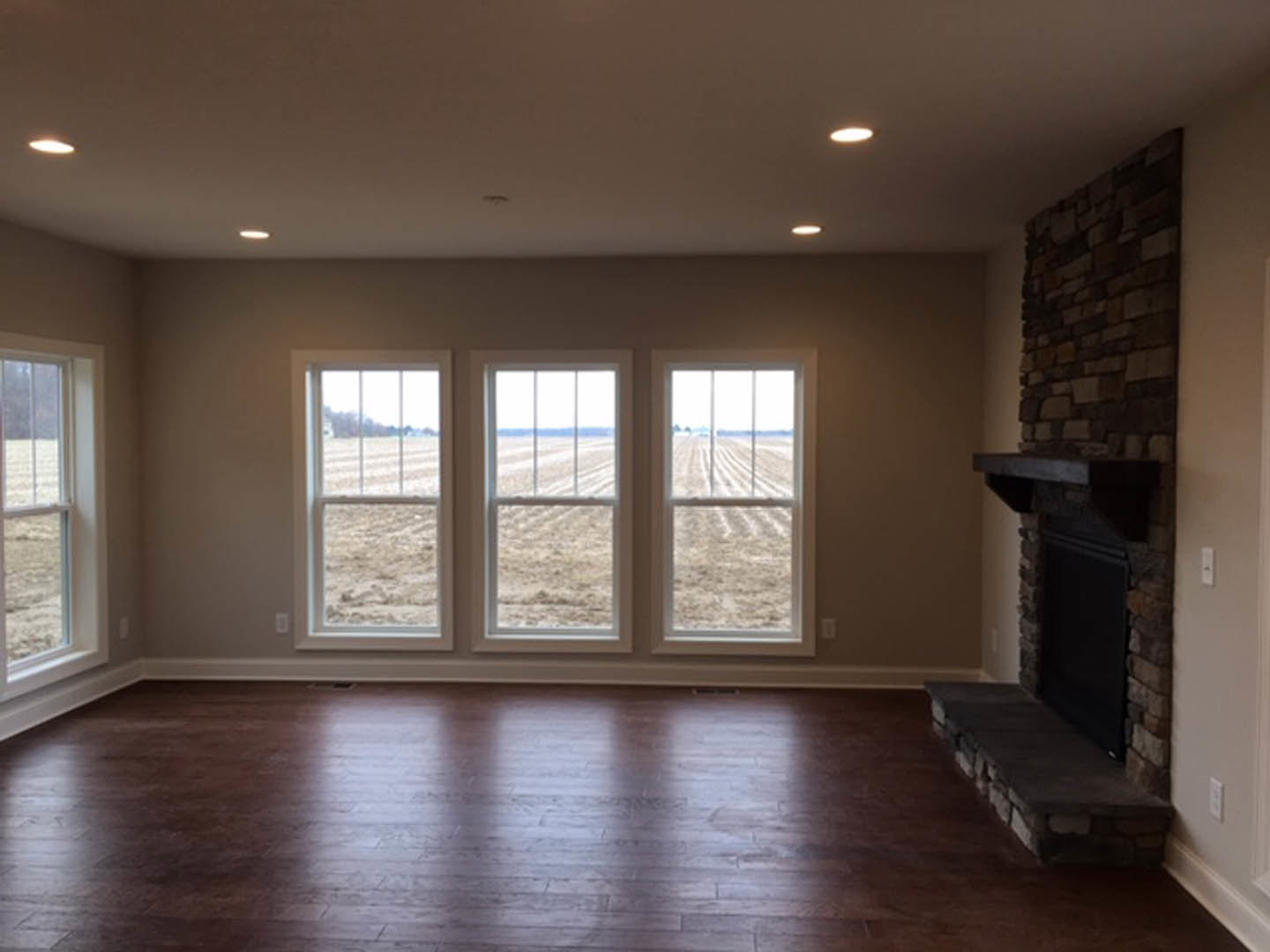 Stone fireplace with raised hearth, row of large windows overlooking a field, wood flooring with natural light reflecting across the surface