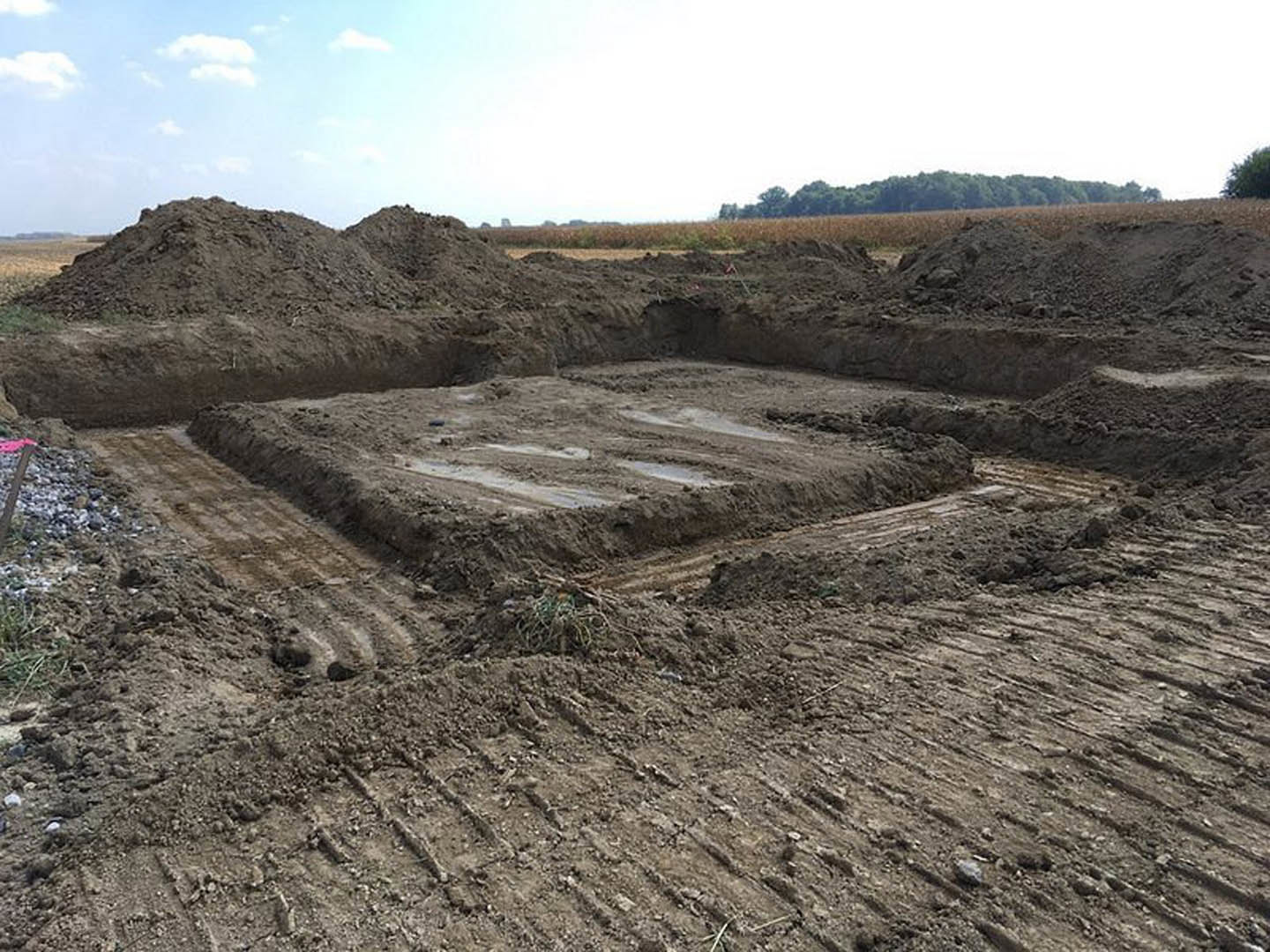 Dirt field with tire tracks and several dirt piles, cloudy white sky overhead, group of trees in the background, blurry hand holding a red object in foreground