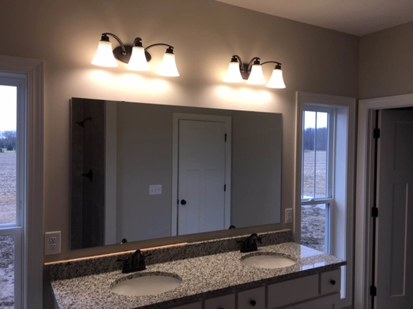 Bathroom with double sinks set in a white countertop, expansive wall mirror above, chrome faucets, light fixture mounted on wall, tiled backsplash, and visible electrical outlet.