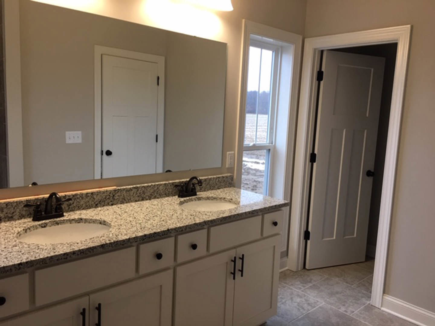 Bathroom with two undermount sinks set in a light stone countertop, wide frameless mirror above, white cabinetry with black hardware, and neutral tile backsplash.