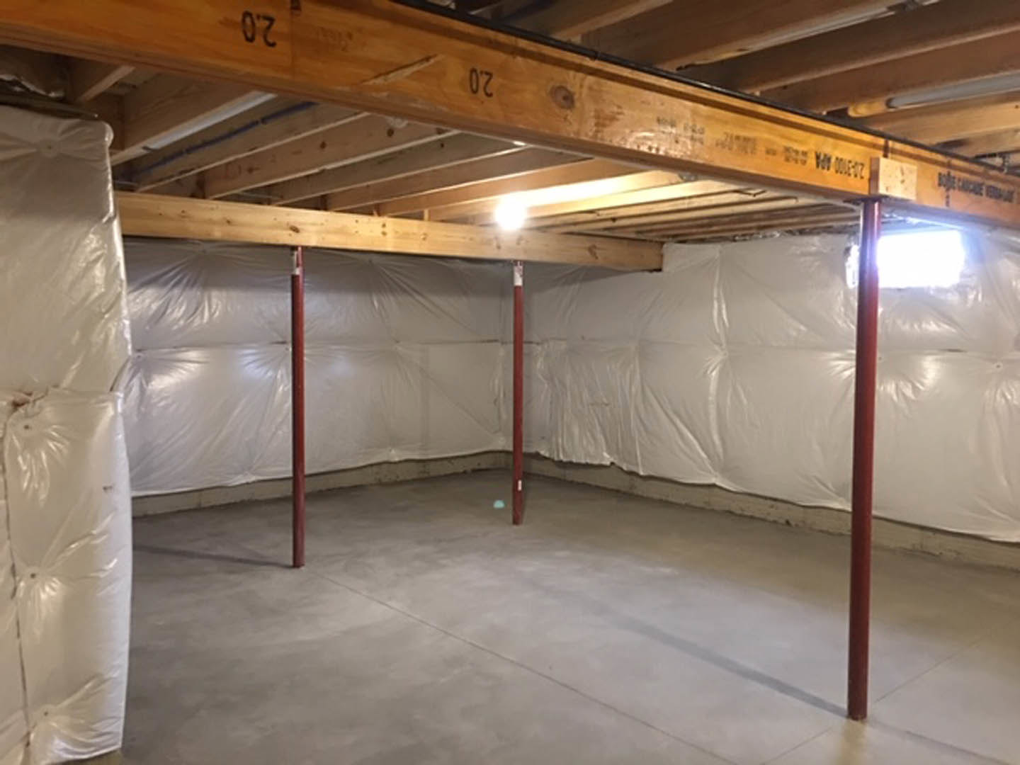 Basement room with exposed wooden ceiling beam, white plastic sheeting covering walls, concrete floor, and metal support pole.