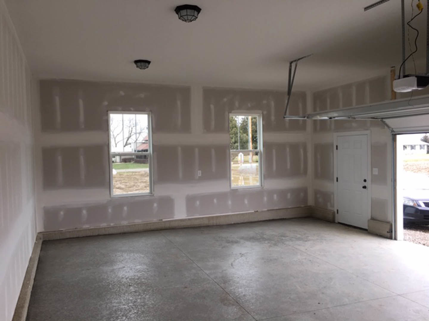 Spacious garage with concrete flooring, white walls, multiple windows offering views of grass and trees, white door with black hardware, ceiling-mounted light fixture