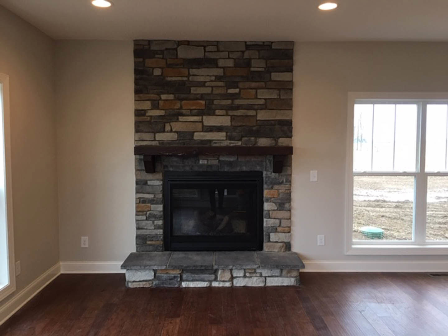 Stone fireplace with wood mantle in a den, hardwood floors, large window offering outdoor view, textured stone wall detailing