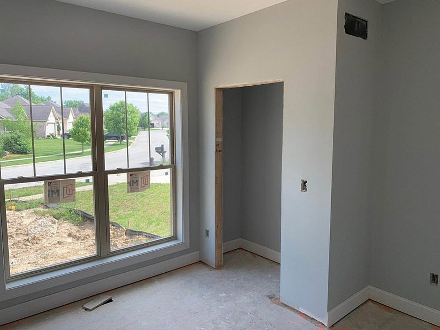 Bright room with white walls, wood flooring, large window overlooking yard and neighboring houses, black rectangular thermostat on wall, white door with wood trim